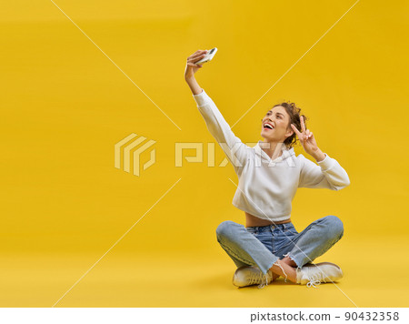Young girl taking selfie with smartphone, sitting with crossed legs in studio. Front view of smiling female showing victory sign, taking selfie photo, isolated on orange background. Concept of selfie. 90432358