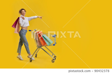 Beautiful female shopaholic pointing away, while shopping in studio. Side view of cute girl with shopping trolley, tiptoeing, looking at camera, isolated on orange background. Concept of shopping. Beautiful female shopaholic pointing away, while shopping in studio. Side view of cute girl with shopping trolley, tiptoeing, looking at camera, isolated on orange background. Concept of shopping. 90432378