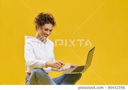 Pretty girl using laptop to make payment online in studio. Front view of smiling woman shopping online, holding plastic card, isolated on orange studio background, copy space. Concept of shopping. Pretty girl using laptop to make payment online in studio. Front view of smiling woman shopping online, holding plastic card, isolated on orange studio background, copy space. Concept of shopping. 90432398