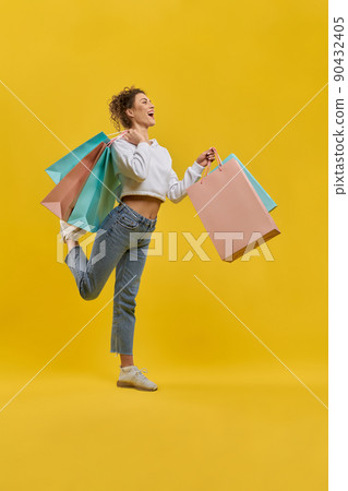 Attractive woman in casual outfit, shopping, smiling inside. Side view of pretty girl with shopping bags, flirty bending one leg on knee, isolated on orange studio background. Concept of shopping.  90432405