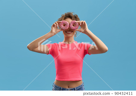 Unrecognizable cheerful woman covering face with colorful donuts inside. Portrait view of woman holding donuts over eyes, smiling at camera, isolated on azure studio background. Concept of amusement. 90432411
