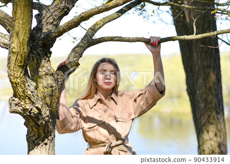 Girl in a jacket near a tree in the forest near the lake Girl in a jacket near a tree in the forest near the lake 90433914