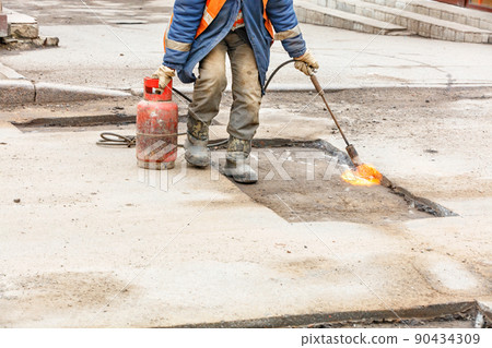 A worker warms up old asphalt with a gas burner on a repaired section of the road. A worker warms up old asphalt with a gas burner on a repaired section of the road. 90434309