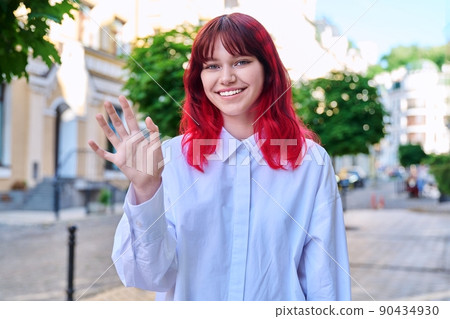 Portrait of smiling teenage female waving hand looking at camera on city street Portrait of smiling teenage female waving hand looking at camera on city street 90434930