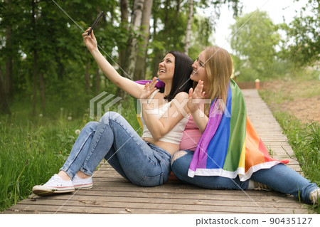 Pride concept. Young happy couple of Caucasian women with LGBT flag communicate via video communication holding phone and waving their hand in sign of welcome while sitting in nature on wooden path Pride concept. Young happy couple of Caucasian women with LGBT flag communicate via video communication holding phone and waving their hand in sign of welcome while sitting in nature on wooden path 90435127