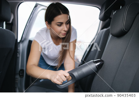 Attractive young woman using portable vacuum cleaner in her car. Woman vacuuming her car in the garage at home. Car interior cleaning 90435403