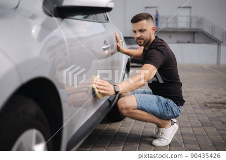 Handsome man wipes a car with a rag in a showroom at a self-service car wash. Gray car. Polished car with hard wax Handsome man wipes a car with a rag in a showroom at a self-service car wash. Gray car. Polished car with hard wax 90435426