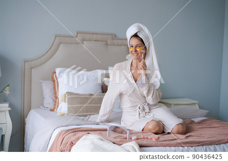 Happy woman sitting in bed, doing facial massage with jade face roller 90436425