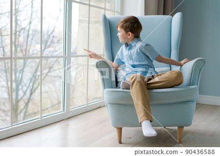 Cute boy sits in chair by window and looks out for something in window Cute boy sits in chair by window and looks out for something in window 90436588