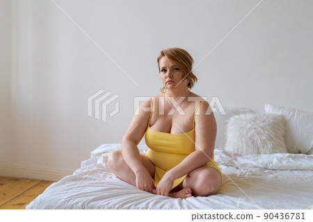 Plus-size model in yellow slinky dress sits on bed beside her bedroom 90436781