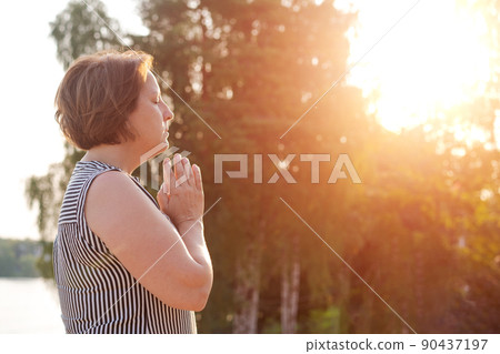 Peaceful woman holds her palms in front her in prayer against backdrop trees Peaceful woman holds her palms in front her in prayer against backdrop trees 90437197