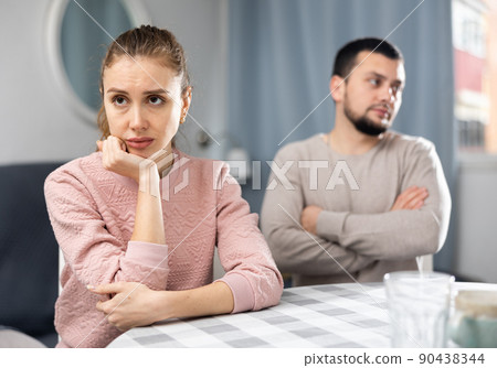 Upset woman sitting at table on background of displeased husband Upset woman sitting at table on background of displeased husband 90438344