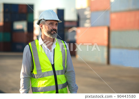 Caucasian male manager in white hard hat helmet and high-visibility vest walking in Container Terminal. Caucasian male manager in white hard hat helmet and high-visibility vest walking in Container Terminal. 90443580