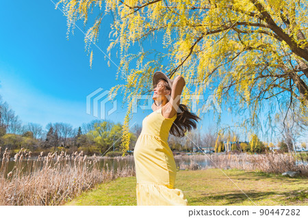 Happy Asian pregnant woman dancing free in nature summer park wearing sun hat and yellow dress. Pregnancy joy and carefree 90447282
