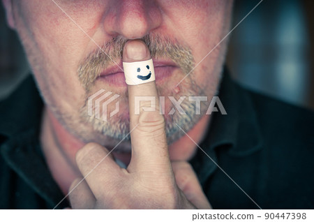 A man shows the gesture of silence with his index finger. A white bandage on the index finger of a grown man's hand. Smiling face painted on the armband. Joy of silence concept. Selective focus. 90447398