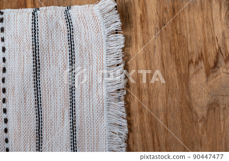A white kitchen towel lies on the countertop of a wooden table. The fringed towel is fringed and patterned on the rough boards. Top view. Copy space A white kitchen towel lies on the countertop of a wooden table. The fringed towel is fringed and patterned on the rough boards. Top view. Copy space 90447477