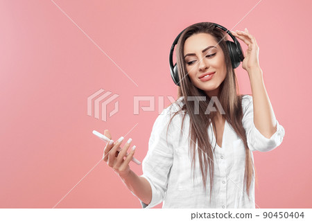 Beautiful woman in headphones with smartphone listening music over pink background. Cute girl uses wireless earphones. Beautiful woman in headphones with smartphone listening music over pink background. Cute girl uses wireless earphones. 90450404