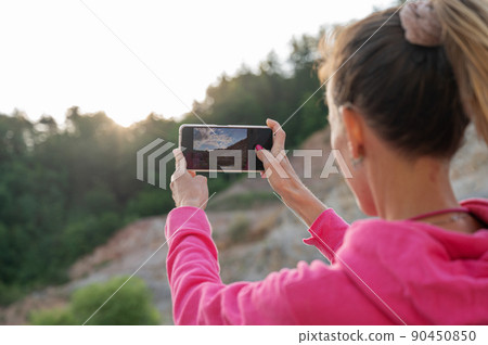 Young woman using her mobile phone to take photos of a sun rising from behind trees Young woman using her mobile phone to take photos of a sun rising from behind trees 90450850