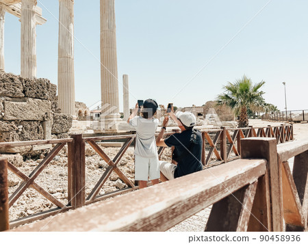 Young father dad and his school boy kid son tourists taking photos of ancient antique coliseum amphitheater ruins on their smartphone, gadget, tablet pc computer in hot summer day 90458936