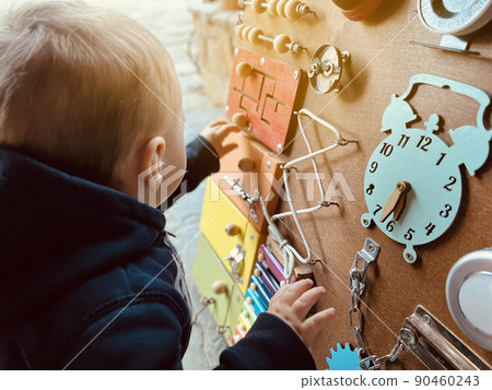 A little boy is playing with a busy board. Developming toy for children from one year old. Developming toy for children from one year old. Focus on the busy board A little boy is playing with a busy board. Developming toy for children from one year old. Developming toy for children from one year old. Focus on the busy board 90460243