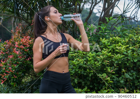Slim sporty girl drinking water. Fitness young woman taking a break after training in park. 90460512