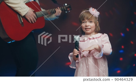 Little girl on stage in vintage dress, she sings into microphone and dances, her father plays acoustic guitar. Color music is shining and smoke is billowing. Father and Child Day, performance on stage Little girl on stage in vintage dress, she sings into microphone and dances, her father plays acoustic guitar. Color music is shining and smoke is billowing. Father and Child Day, performance on stage 90464329