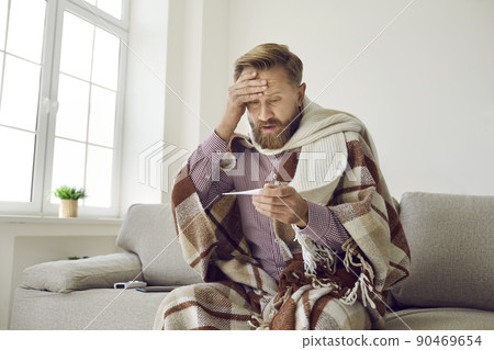 Sick man who is afraid of spread of coronavirus looks scared at thermometer shows high temperature. 90469654