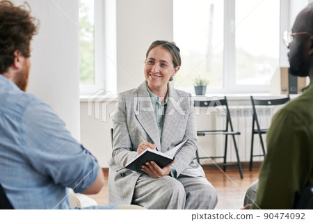 Happy female psychologist making notes in notebook while listening to patient 90474092