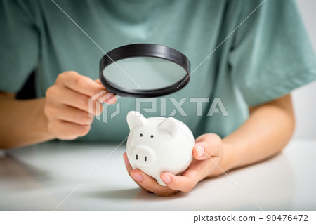 Young woman with magnifying glass looking at the piggy bank. Financial audit concept. 90476472