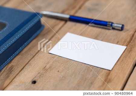 Blue diary, business card and pencil opposite wooden background. Closed hardcover notebook. White blank business card. Metallic silver-blue pencil rests on top of old cracked boards. Selective focus 90480964