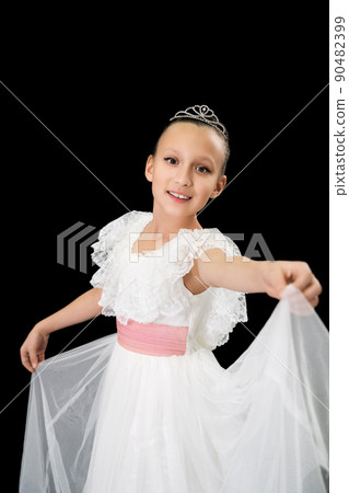 Smiling girl ballet dancer with diadem on head dressed in white long dress dancing on black background in studio. Caucasian ballerina nine-year-old. Part of photo series 90482399