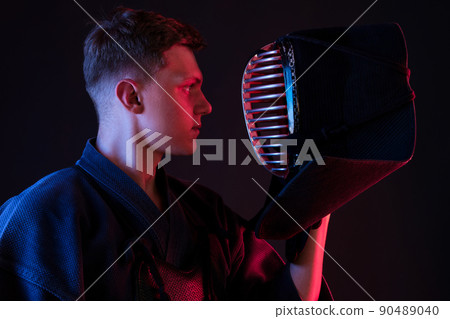 Close up shot, Kendo fighter wearing in an armor, traditional kimono is looking at his helmet while standing against a black studio background. 90489040
