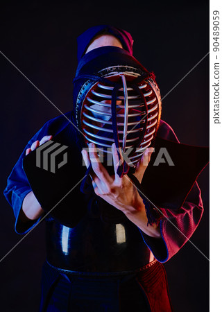 Close up shot, Kendo fighter wearing in an armor and traditional kimono is tying the lacing on his helmet standing against a black background. 90489059