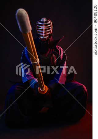 Close up. Kendo fighter wearing in an armor, traditional kimono, helmet, sitting, practicing martial art with shinai bamboo sword, black background. Close up. Kendo fighter wearing in an armor, traditional kimono, helmet, sitting, practicing martial art with shinai bamboo sword, black background. 90489120