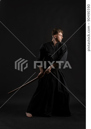 Kendo guru wearing in a traditional japanese kimono is practicing martial art with the shinai bamboo sword against a black studio background. 90490390