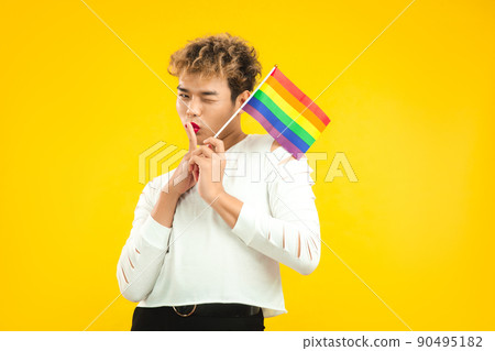 Young asian pretty gay man in white t-shirt holding lgbt pride flag posing on the yellow color background. Young asian pretty gay man in white t-shirt holding lgbt pride flag posing on the yellow color background. 90495182
