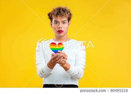 Hopeful Young asian gay man holding rainbow paper cut heart in concept of lgbtq and lgbtq+ posing isolated yellow background. 90495228