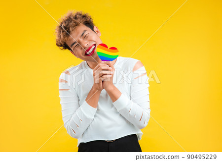 Happy Young asian gay man holding rainbow paper cut heart in concept of lgbtq and lgbtq+ posing isolated yellow color background. 90495229