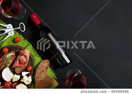 Prosciutto, salami, baguette slices, tomatoes and nutson rustic wooden board, two glasses of red wine over black background Prosciutto, salami, baguette slices, tomatoes and nutson rustic wooden board, two glasses of red wine over black background 90498406