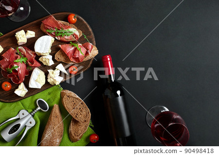 Delicious appetizer to wine - ham, cheese, baguette slices, tomatoes, served on a wooden board, and glass with red wine on black surface 90498418