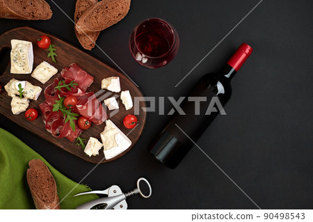 Red wine, cheese, cherry tomato, bread and prosciutto on wooden board over black backdrop, top view, copy space 90498543