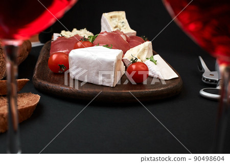 Prosciutto, salami, baguette slices, tomatoes and nutson rustic wooden board, two glasses of red wine over black background Prosciutto, salami, baguette slices, tomatoes and nutson rustic wooden board, two glasses of red wine over black background 90498604