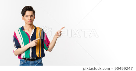 Skeptical and displeased young man having concern, poitning fingers right and looking with disapproval, standing over white background 90499247