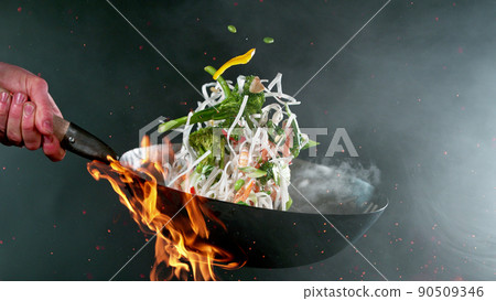 Closeup of chef throwing vegetable noodle mix from wok pan in fire. Closeup of chef throwing vegetable noodle mix from wok pan in fire. 90509346