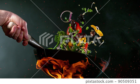Closeup of chef throwing vegetable mix from wok pan in fire. 90509351
