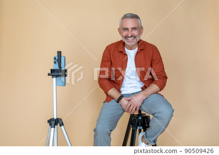 A man in a red jacket sitting on a stool in a studio and making selfie A man in a red jacket sitting on a stool in a studio and making selfie 90509426