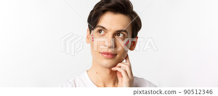 People, lgbtq and beauty concept. Close-up of happy queer man with applied lip gloss and glitter, smiling and looking at upper left corner, standing over white background 90512346