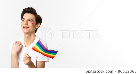 Waist-up shot of attractive gay man with glitter on face, waving pride rainbow flag and looking dreamy at upper left corner, lgbtq concept 90512363