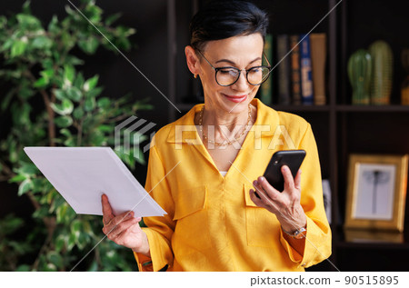 Middle aged businesswoman using smartphone and reading data on paper 90515895