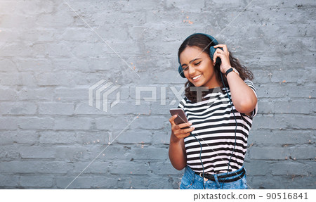 Getting her weekend playlist together. Shot of a young woman using a cellphone while wearing headphones against a grey wall. 90516841
